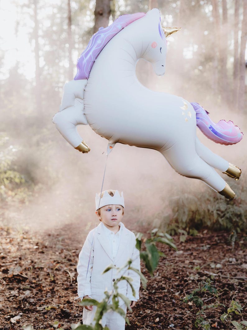 May include: A young boy wearing a white crown and a white suit stands in a woodland setting with a large white unicorn balloon floating above him.