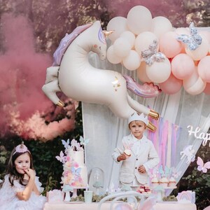 May include: A unicorn balloon with gold accents and a pink and purple colour scheme, a child wearing a white suit and a crown, and a cake decorated with butterflies and flowers. The scene is set up for a birthday party with a unicorn theme.