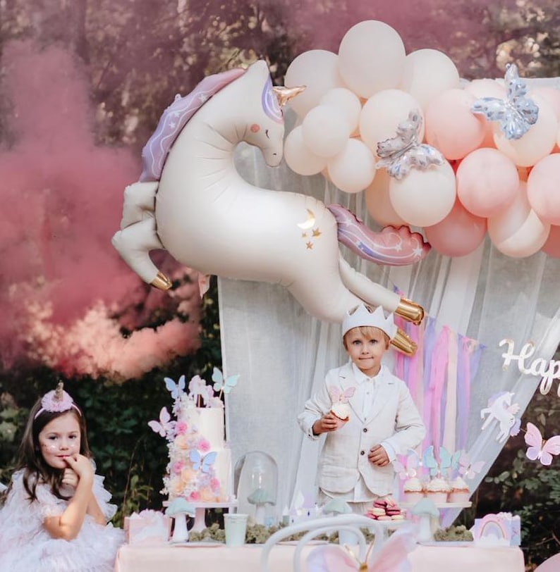 May include: A unicorn balloon with gold accents and a pink and purple colour scheme, a child wearing a white suit and a crown, and a cake decorated with butterflies and flowers. The scene is set up for a birthday party with a unicorn theme.
