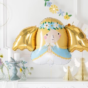 A gold and pastel-colored foil balloon shaped like a praying angel with golden wings, blonde hair, and a floral crown. The balloon is part of a soft, elegant party setup with pastel flowers, candles, and paper angel decorations in the background, perfect for a baptism, communion, or baby celebration.
