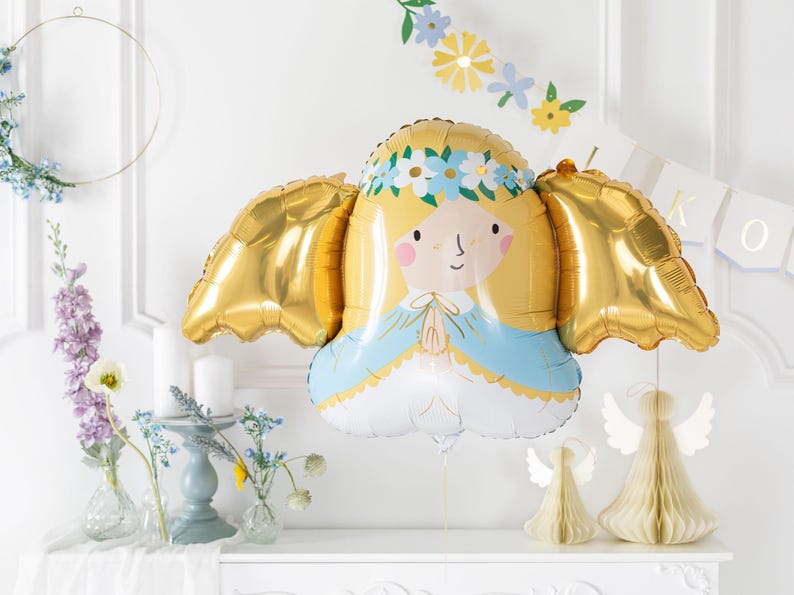 A gold and pastel-colored foil balloon shaped like a praying angel with golden wings, blonde hair, and a floral crown. The balloon is part of a soft, elegant party setup with pastel flowers, candles, and paper angel decorations in the background, perfect for a baptism, communion, or baby celebration.