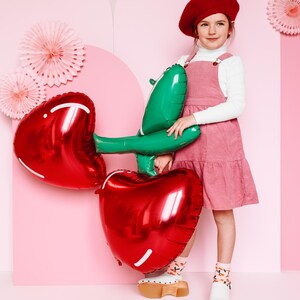May include: A girl wearing a pink dress and a red beret holds two large red heart-shaped balloons with green stems. The balloons are shaped like cherries.