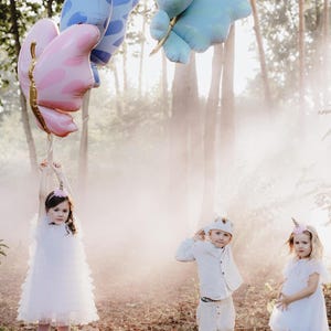 Pu&ograve; includere: Tre bambini vestiti di bianco e con fasce per capelli a forma di unicorno e corona posano in un ambiente boschivo nebbioso. Tengono palloncini a forma di farfalla colorati.