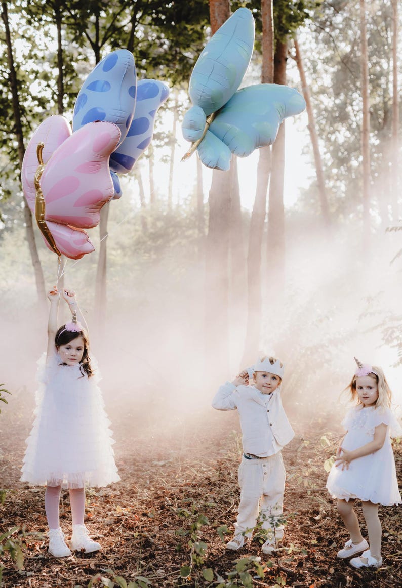 Pu&ograve; includere: Tre bambini vestiti di bianco e con fasce per capelli a forma di unicorno e corona posano in un ambiente boschivo nebbioso. Tengono palloncini a forma di farfalla colorati.