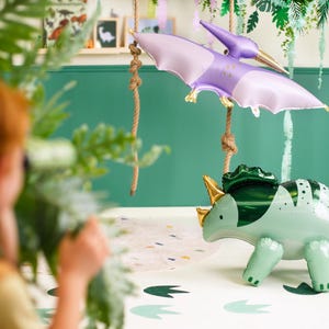 Puede incluir: Un globo de dinosaurio verde y dorado con un globo de pterod&aacute;ctilo morado colgando sobre &eacute;l. Los globos est&aacute;n frente a una pared verde con serpentinas verdes y blancas colgando del techo.