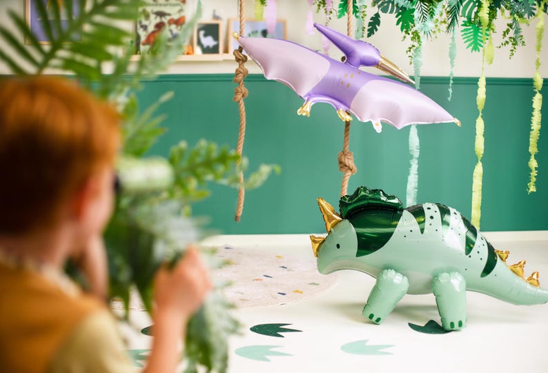 Puede incluir: Un globo de dinosaurio verde y dorado con un globo de pterod&aacute;ctilo morado colgando sobre &eacute;l. Los globos est&aacute;n frente a una pared verde con serpentinas verdes y blancas colgando del techo.