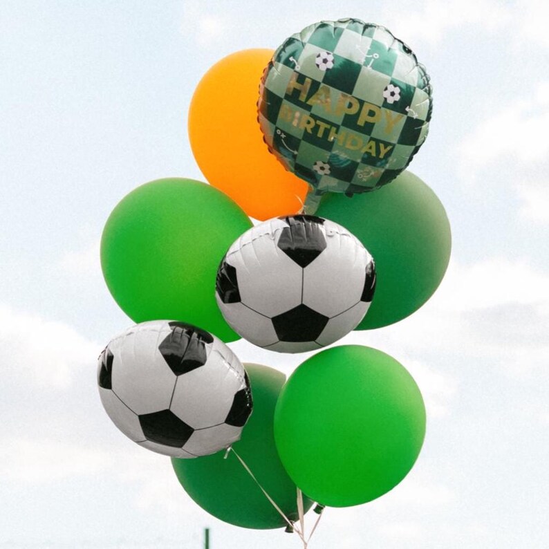 May include: A cluster of colorful balloons against a light blue sky. The arrangement includes green, orange, and soccer ball-themed balloons. One balloon reads "HAPPY BIRTHDAY" in gold lettering. Perfect for a birthday celebration.