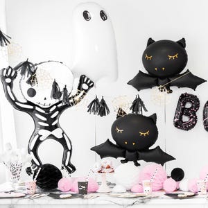 May include: Halloween party decorations featuring a skeleton, ghost, and bat-shaped balloons. The black and white balloons are accented with gold spiderwebs and tassels. Pink and black party supplies are arranged on a table.