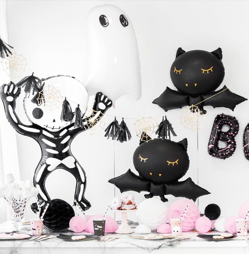 May include: Halloween party decorations featuring a skeleton, ghost, and bat-shaped balloons. The black and white balloons are accented with gold spiderwebs and tassels. Pink and black party supplies are arranged on a table.