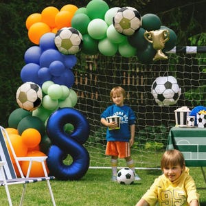 May include: A festive outdoor scene with a soccer theme. A balloon arch in orange, green, blue, and soccer ball designs frames a soccer net. A large blue number 8 balloon is present. Two children are in the scene, one holding a box of popcorn.