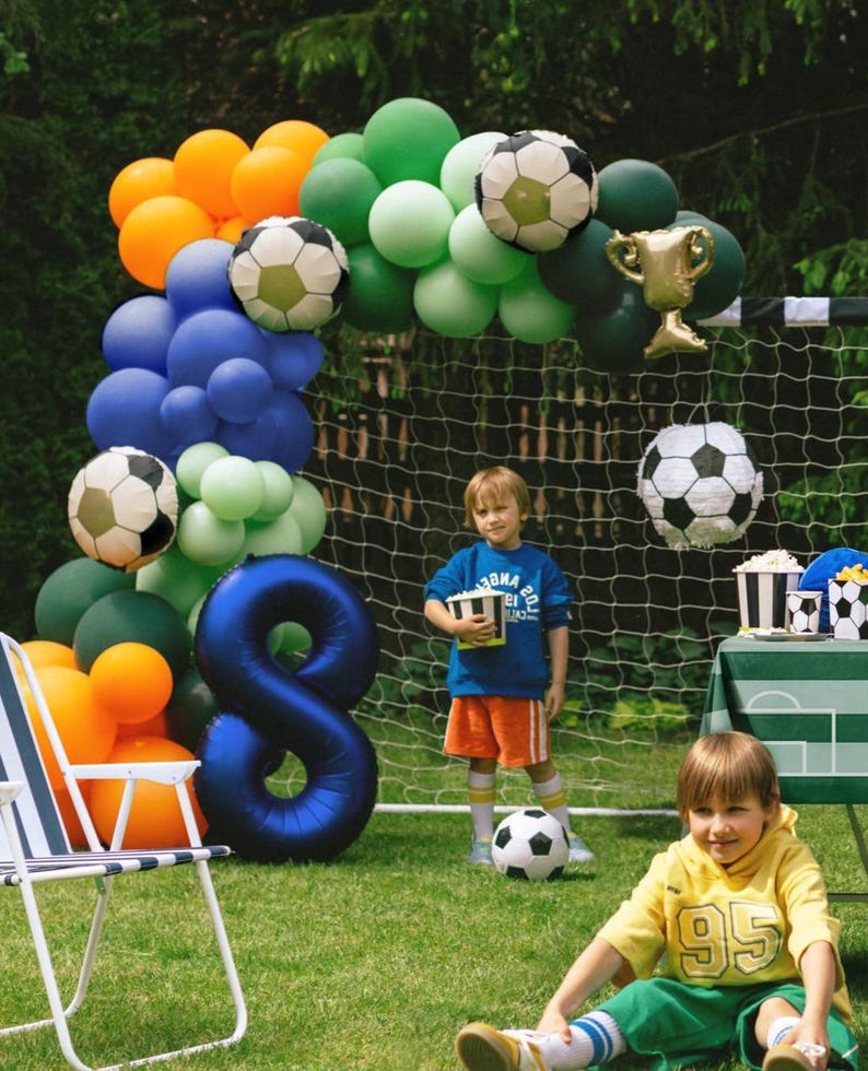 May include: A festive outdoor scene with a soccer theme. A balloon arch in orange, green, blue, and soccer ball designs frames a soccer net. A large blue number 8 balloon is present. Two children are in the scene, one holding a box of popcorn.