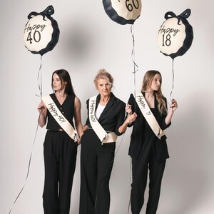 May include: Three women wearing black outfits and sashes with the text "Happy 40", "Happy 60", and "Happy 18" stand in front of a white background. They are holding black and white balloons with the same text as their sashes.