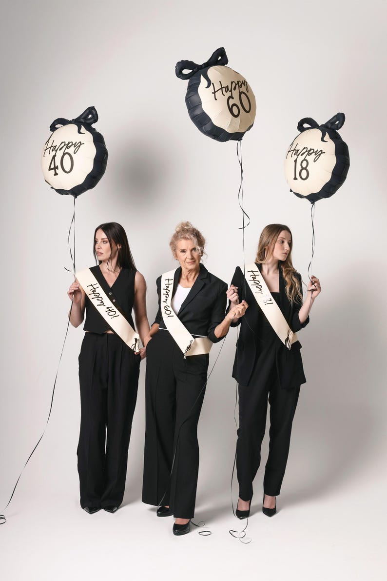 May include: Three women wearing black outfits and sashes with the text "Happy 40", "Happy 60", and "Happy 18" stand in front of a white background. They are holding black and white balloons with the same text as their sashes.