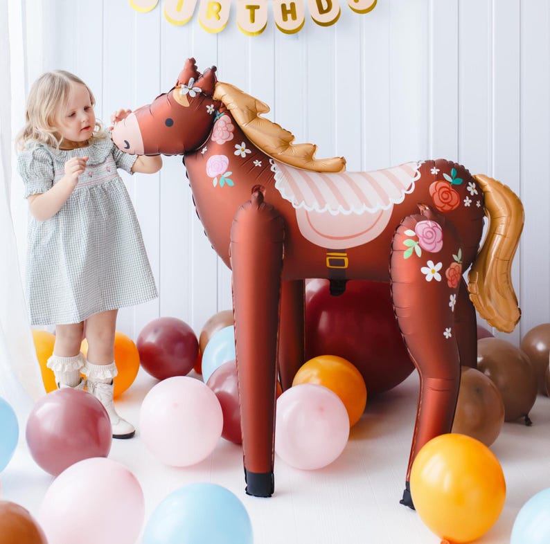 May include: A brown horse balloon with a pink and white saddle and floral details. The balloon is surrounded by colorful balloons and a young girl is touching the horse's nose.