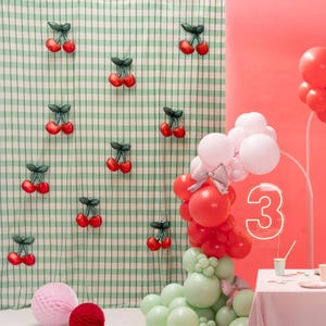 K&ouml;nnte beinhalten: Eine festliche Party-Szene mit roten und gr&uuml;nen Luftballons, einer rosa Wabenkugel und kirschf&ouml;rmigen Dekorationen auf einem gr&uuml;n-wei&szlig; karierten Hintergrund. Eine Neon-Zahl 3 ist ebenfalls zu sehen.