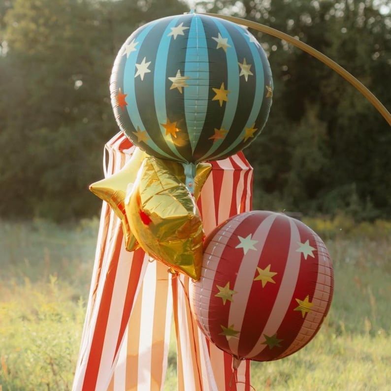 May include: A cluster of festive balloons in a grassy outdoor setting. The balloons include a large blue and black striped sphere with gold stars, a gold star-shaped balloon, and a red and white striped sphere with gold stars.