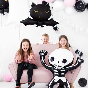 以下が含まれることがあります： ピンクのソファに3人の子供が座っています。大きな黒と白のスケルトンバルーンがあります。子供たちはハロウィンのコスチュームを着ており、背景には黒とピンクのバルーンがあります。