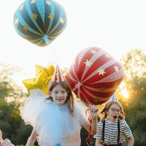 May include: Children celebrating outdoors with colorful balloons. One balloon is blue with gold stars, another is red with white stripes and gold stars. The children wear party hats and costumes, creating a festive atmosphere.