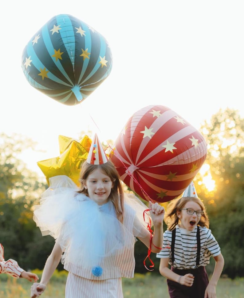 May include: Children celebrating outdoors with colorful balloons. One balloon is blue with gold stars, another is red with white stripes and gold stars. The children wear party hats and costumes, creating a festive atmosphere.
