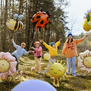 May include: Five children in a woodland setting holding colourful balloons shaped like a ladybug, bee, sun, and flowers. The balloons have happy faces and some have text on them, including "I'll be working til I'm full".