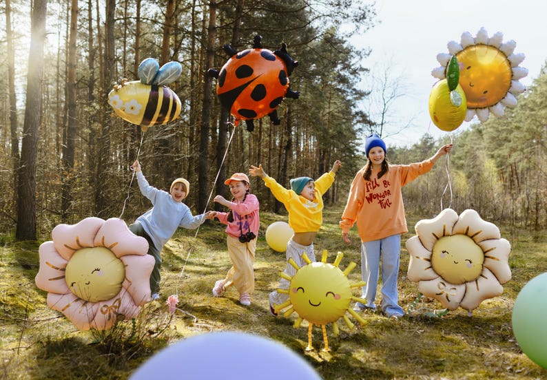 May include: Five children in a woodland setting holding colourful balloons shaped like a ladybug, bee, sun, and flowers. The balloons have happy faces and some have text on them, including "I'll be working til I'm full".