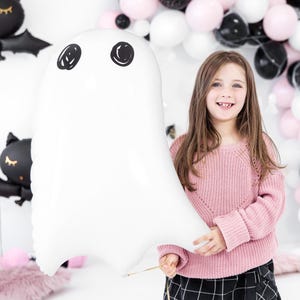 May include: A white ghost-shaped balloon with black eyes is held by a person. The balloon is the focal point, with a gold string attached. The background includes a variety of balloons in black, white, and pink, suggesting a Halloween or party theme.