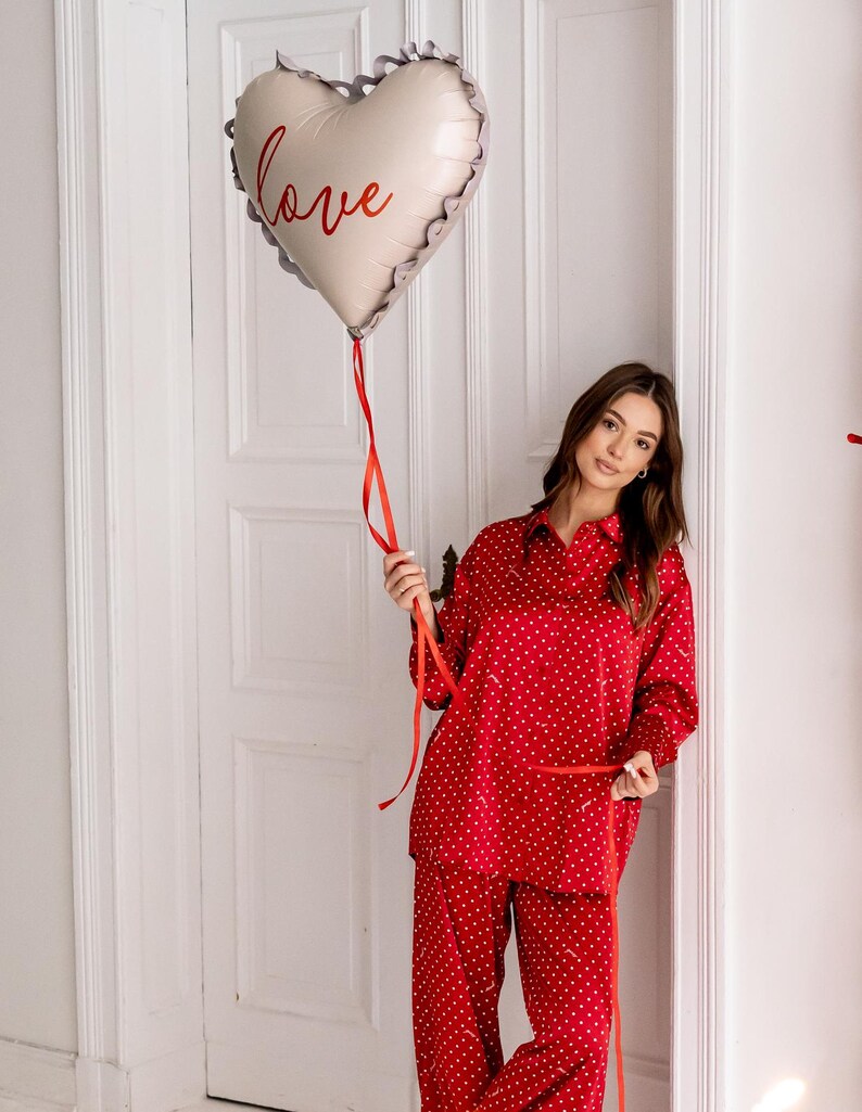Woman in a red polka dot pyjamas holding a heart-shaped foil balloon with the word "love" written in red script. The balloon has a ruffled edge and a red ribbon, and she is standing in front of a white paneled wall, creating a romantic and elegant atmosphere.