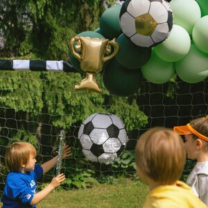 May include: A soccer-themed party scene with a soccer ball pi&ntilde;ata, a gold trophy balloon, and soccer ball balloons. Children are playing in front of a soccer net, with green and white balloons in the background.