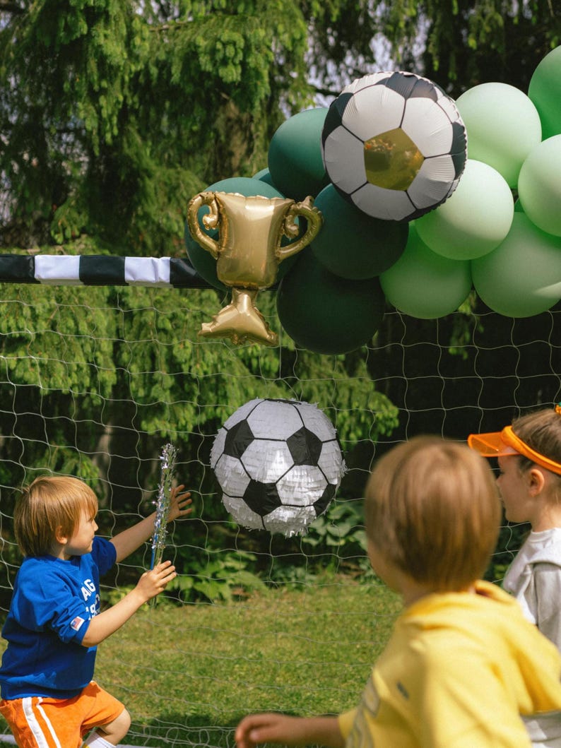 May include: A soccer-themed party scene with a soccer ball pi&ntilde;ata, a gold trophy balloon, and soccer ball balloons. Children are playing in front of a soccer net, with green and white balloons in the background.