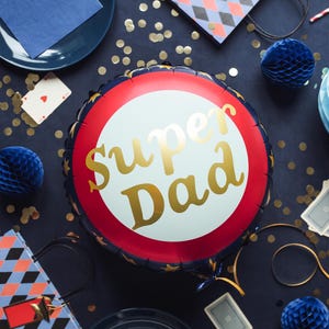 May include: A round foil balloon with the words "Super Dad" in gold script. The balloon has a white center, a red ring, and a navy blue border with gold stars. Party decorations, including blue honeycomb balls and playing cards, surround the balloon.