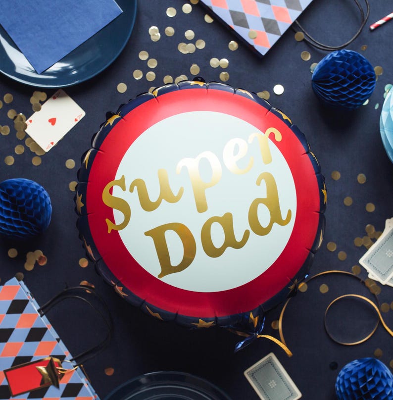 May include: A round foil balloon with the words "Super Dad" in gold script. The balloon has a white center, a red ring, and a navy blue border with gold stars. Party decorations, including blue honeycomb balls and playing cards, surround the balloon.