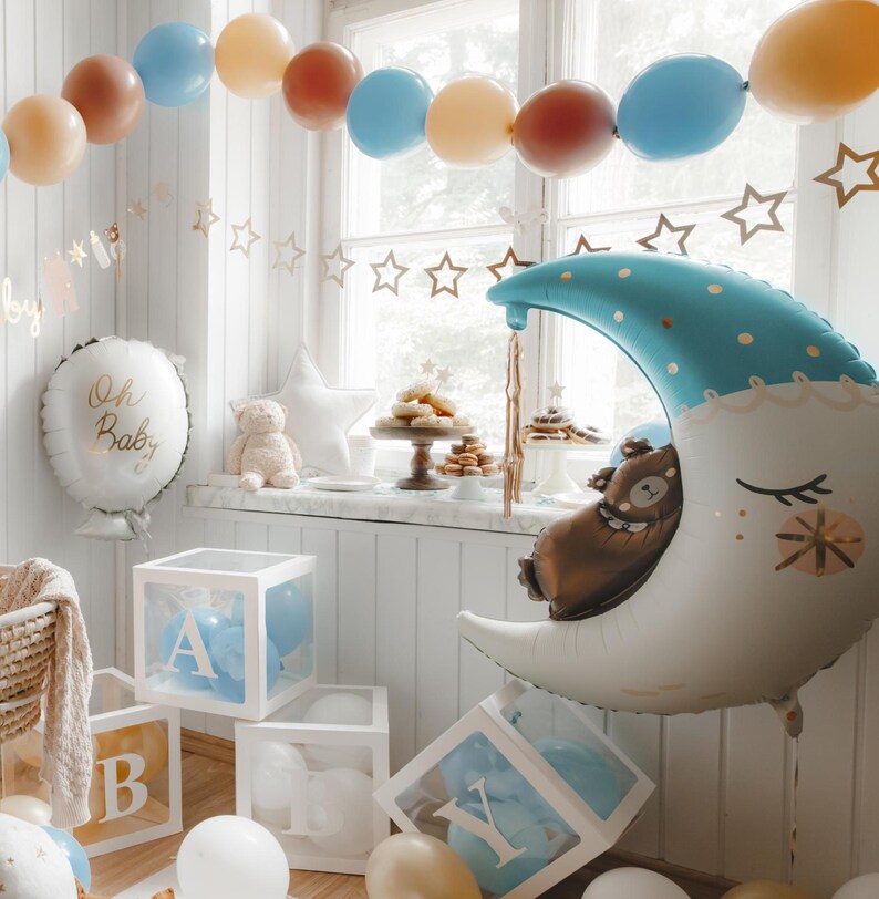 May include: A baby shower decoration with a blue and white moon balloon, a white balloon with the text "Oh Baby", and a table with a cake and other treats. There are also blue and brown balloons in the background.