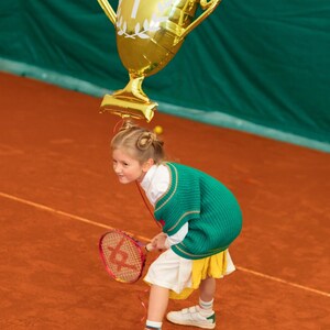 Puede incluir: Una ni&ntilde;a peque&ntilde;a con un su&eacute;ter verde y una falda blanca sostiene una raqueta de tenis y un globo dorado con forma de trofeo con el n&uacute;mero "1" en &eacute;l. La ni&ntilde;a est&aacute; de pie en una cancha de tenis de tierra batida roja.