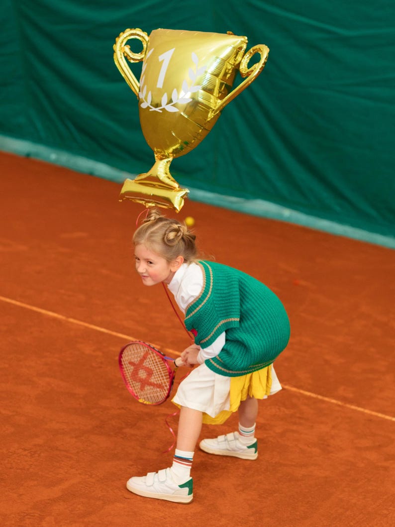 Puede incluir: Una ni&ntilde;a peque&ntilde;a con un su&eacute;ter verde y una falda blanca sostiene una raqueta de tenis y un globo dorado con forma de trofeo con el n&uacute;mero "1" en &eacute;l. La ni&ntilde;a est&aacute; de pie en una cancha de tenis de tierra batida roja.