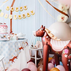 May include: A brown and white horse balloon with floral details and a pink and white striped saddle. The balloon is surrounded by pink, brown, and orange balloons. A table with a blue and white checkered tablecloth is set with a cake and other decorations. A banner with the words "Happy Birthday" hangs above the table.