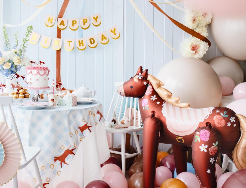 May include: A brown and white horse balloon with floral details and a pink and white striped saddle. The balloon is surrounded by pink, brown, and orange balloons. A table with a blue and white checkered tablecloth is set with a cake and other decorations. A banner with the words "Happy Birthday" hangs above the table.