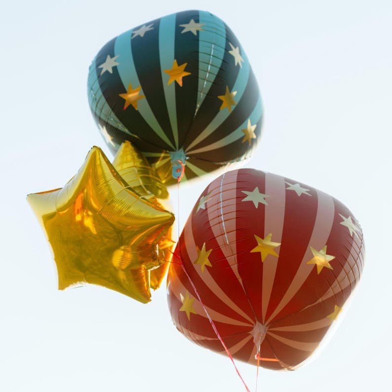 K&ouml;nnte beinhalten: Drei Heliumballons vor hellem Himmel. Einer hat die Form eines goldenen Sterns, die anderen beiden sind quadratisch mit Stern- und Streifenmustern. Ein quadratischer Ballon ist schwarz und blaugr&uuml;n mit goldenen Sternen, der andere ist rot und wei&szlig; mit goldenen Sternen.