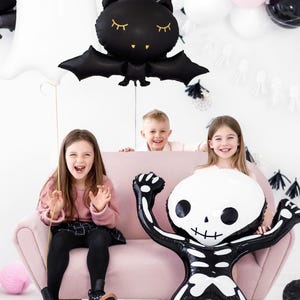 May include: A Halloween-themed scene with children and balloons. A large skeleton balloon is held by a child, while a ghost and bat balloon float above. Pink, black, and white balloons decorate the background. The children are sitting on a pink couch.