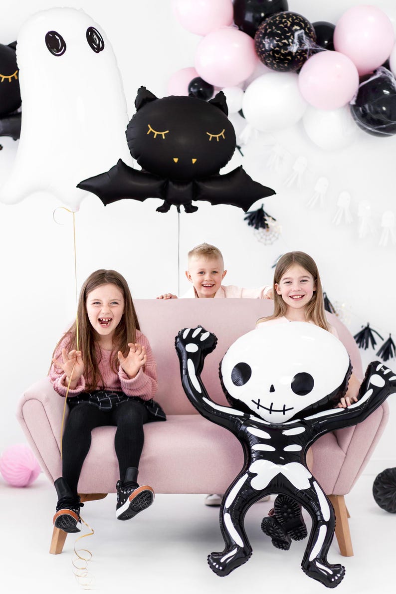 May include: A Halloween-themed scene with children and balloons. A large skeleton balloon is held by a child, while a ghost and bat balloon float above. Pink, black, and white balloons decorate the background. The children are sitting on a pink couch.