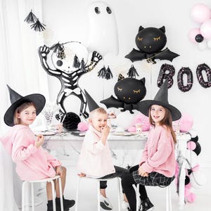 May include: A Halloween-themed party scene with children and decorations. Black bat and ghost balloons, a skeleton balloon, and the word "BOO" in balloon letters are visible. Children wear witch hats and pink clothing.