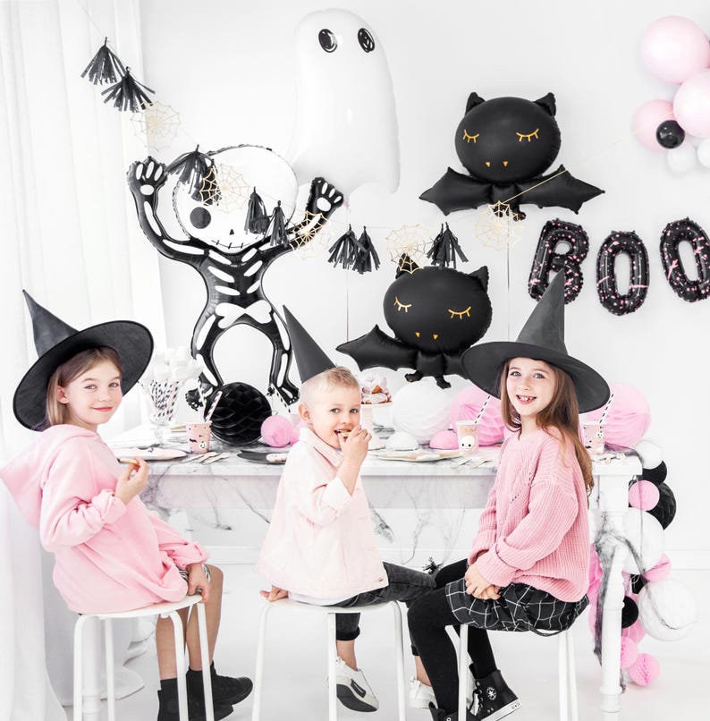 May include: A Halloween-themed party scene with children and decorations. Black bat and ghost balloons, a skeleton balloon, and the word "BOO" in balloon letters are visible. Children wear witch hats and pink clothing.