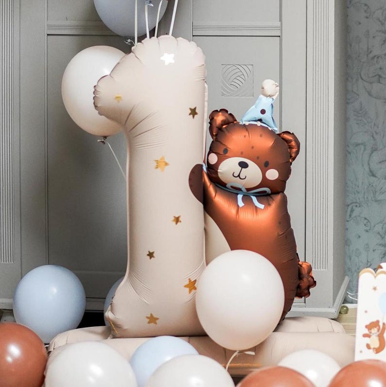 May include: A brown bear balloon wearing a blue party hat, next to a large beige number one balloon with gold stars. There are several other balloons in the background, including white, beige, and blue.