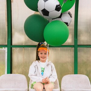 May include: A young person sits on a bench, holding a bundle of balloons. The balloons include green, orange, and soccer ball-themed designs, with one reading "Happy Birthday." The person wears a white jacket, green shorts, and a visor.