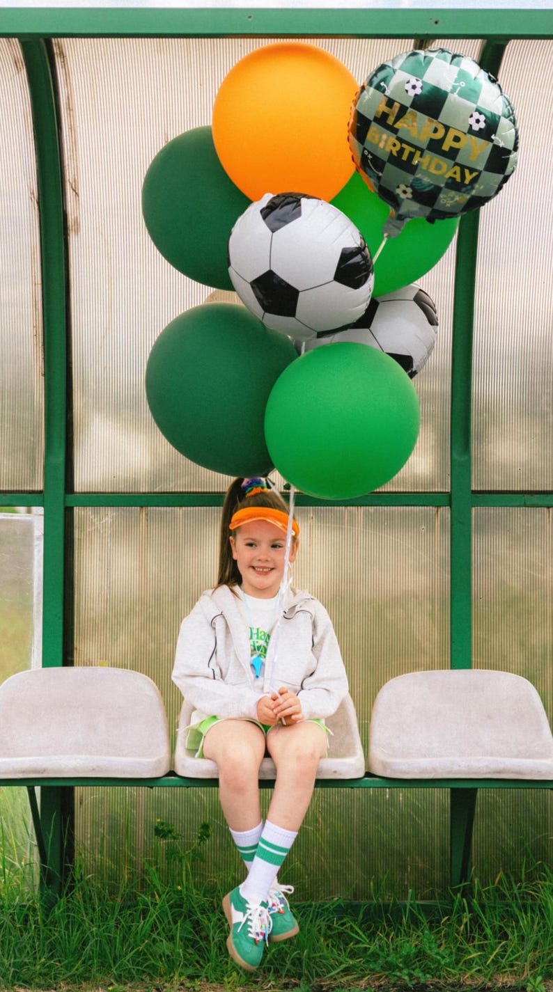 May include: A young person sits on a bench, holding a bundle of balloons. The balloons include green, orange, and soccer ball-themed designs, with one reading "Happy Birthday." The person wears a white jacket, green shorts, and a visor.