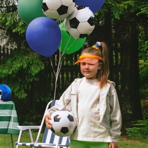 May include: A young person holding a soccer ball, surrounded by colorful balloons. The balloons include soccer ball designs and a "Happy Birthday" balloon. The person is wearing a white jacket, green shorts, and white socks with green stripes.