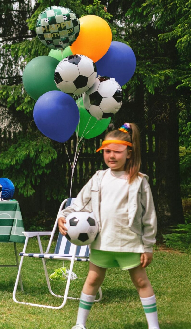 May include: A young person holding a soccer ball, surrounded by colorful balloons. The balloons include soccer ball designs and a "Happy Birthday" balloon. The person is wearing a white jacket, green shorts, and white socks with green stripes.