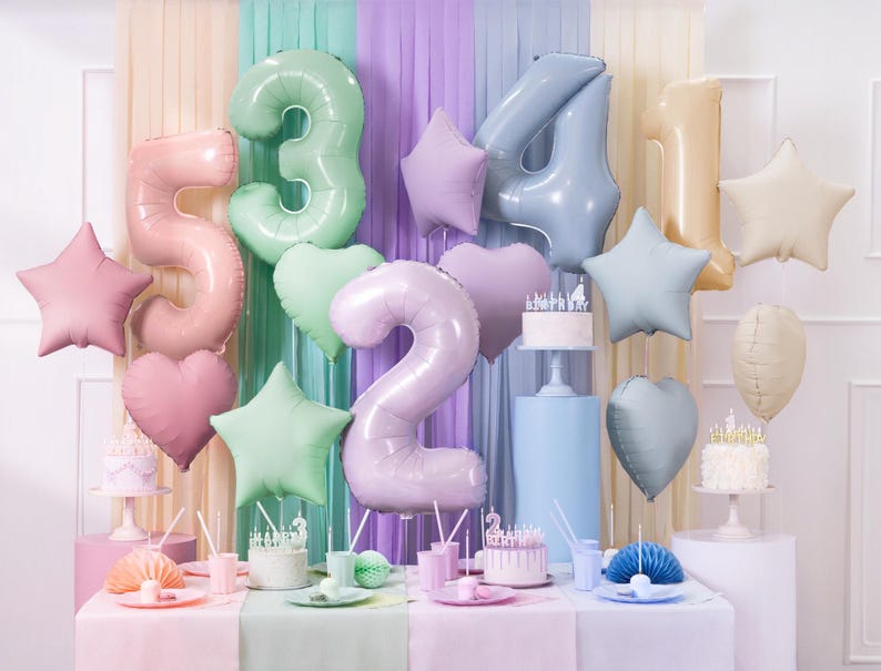 May include: Pastel colored foil balloons in the shape of stars, hearts, and numbers, including 2, 3, 4, and 5. The balloons are hanging from a backdrop of pastel colored fabric. There are two cakes with candles on a table with pastel colored tablecloths.