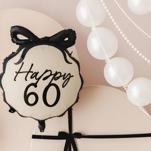 May include: A white and black balloon with a black bow and the text "Happy 60" on it. The balloon is on a light pink surface with a white pearl garland and a black ribbon.