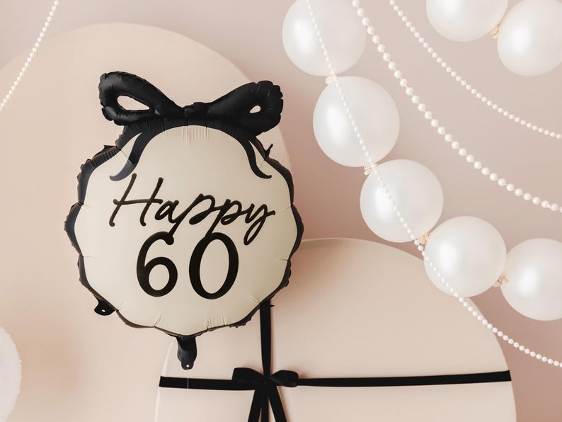 May include: A white and black balloon with a black bow and the text "Happy 60" on it. The balloon is on a light pink surface with a white pearl garland and a black ribbon.