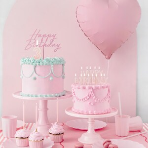 May include: Two pink cakes with pink candles on cake stands. The cakes are decorated with pink frosting and white accents. A pink heart-shaped balloon is in the background. The text "Happy Birthday" is on a pink cake topper.